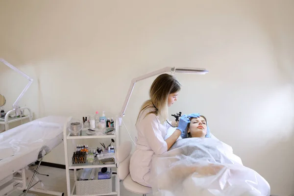 Young woman at cosmetology cabinet getting tattooing eyebrows by cosmetologist.