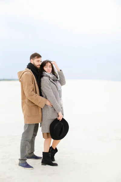 若者は 立っていると白い冬背景の灰色のコートやマフラーを身に着けている女性を抱き締めます ロマンチックなカップルのフォト セッションと季節のインスピレーションの概念 — ストック写真