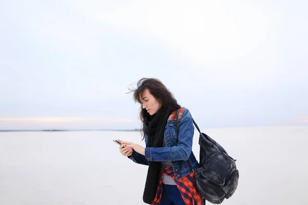 Joven mujer usando jeans y bolso, usando smartphone y de pie en fondo blanco de invierno . —  Fotos de Stock