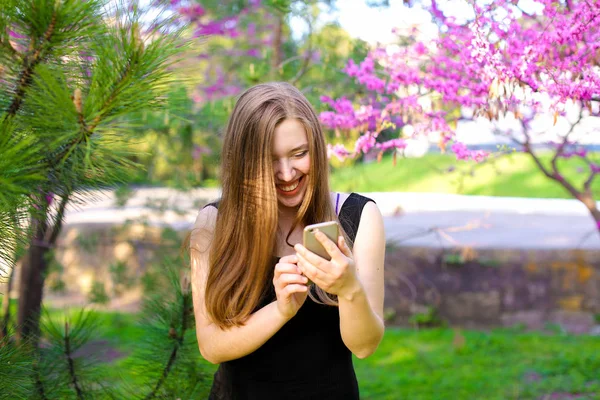 Mujer alegre usando nuevo teléfono inteligente en el parque con los árboles en flor de fondo . — Foto de Stock
