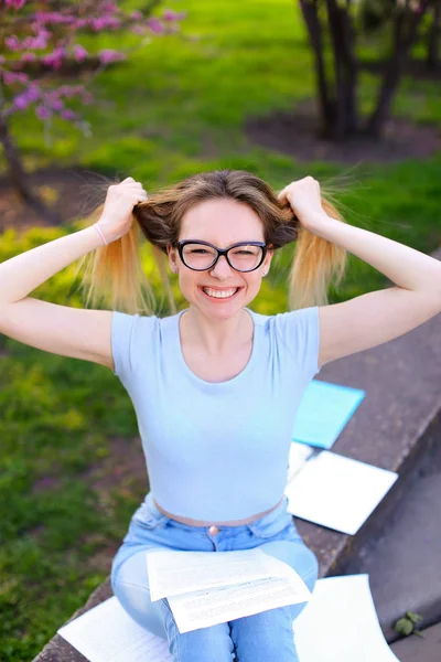 Usmívající se žena studentů čtoucích noviny a hrát si s vlasy na jaře parku. — Stock fotografie