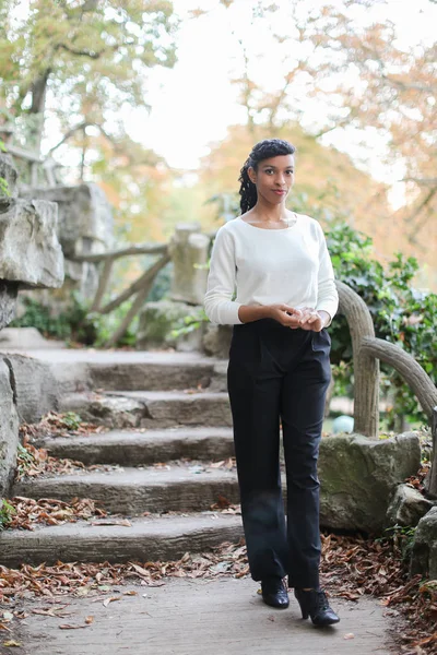 Pretty black girl wearing white blouse standing near stone stairs in park.