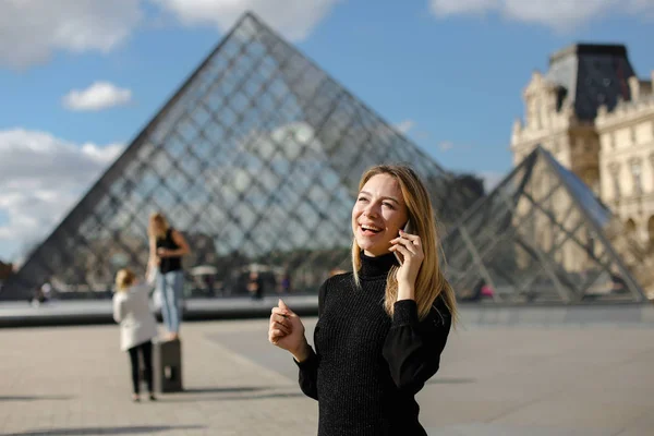 Dívka stojící vedle Louvre a sklo pyramind v černých šatech v Paříži, mluví o smartphone. — Stock fotografie