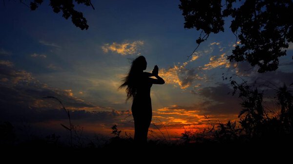 Dark female silhouette doing yoga and meditating in evening sky background. Concept of relaxing before sleeping and asanas.