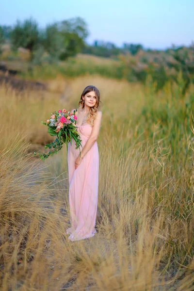 Pregnant woman wearing pink dress and bouquet of flowers standing in steppe background. — Stock Photo, Image
