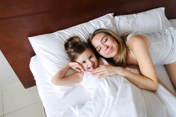 Счастливая мать-блондинка, лежащая в постели с маленькой веселой дочкой . — стоковое фото