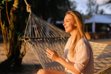 Smartphone kullanarak ve hasır hamak tropikal resort üzerinde oturan genç gülümseyen kadın.