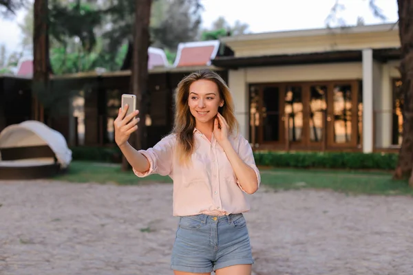 Young caucasian woman making selfie by smartphone outside, house in background.