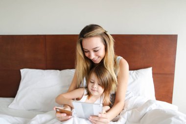 Genç mutlu anne ile küçük kızı yatakta oturuyor ve tablet kullanma.