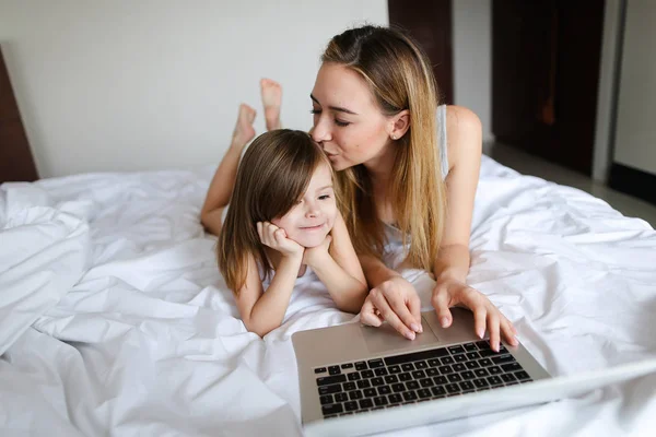 Jovem mulher bonita usando laptop e beijando pequena filha e deitada na cama . — Fotografia de Stock