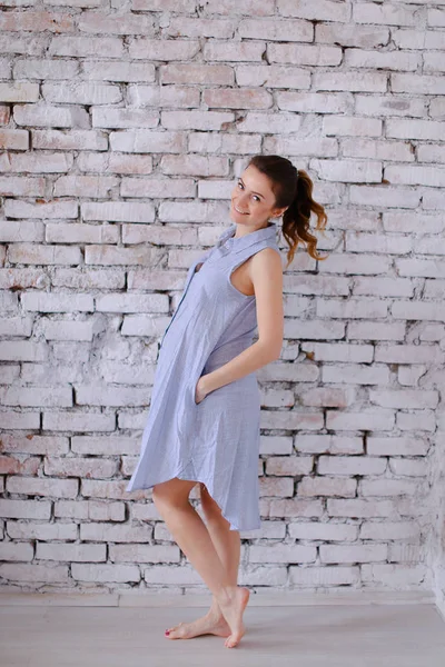 Mujer joven embarazada con vestido azul de pie en el fondo de la pared de ladrillo y la celebración del vientre . —  Fotos de Stock