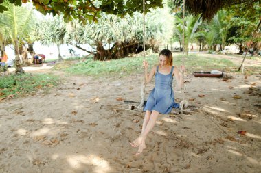 Genç güzel salıncağa binmek ve kot sundress, arka planda kum giyen kız.