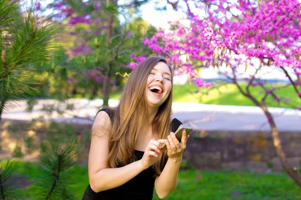 Jocund Mädchen mit neuem Smartphone in Park mit blühenden Bäumen Hintergrund. — Stockfoto