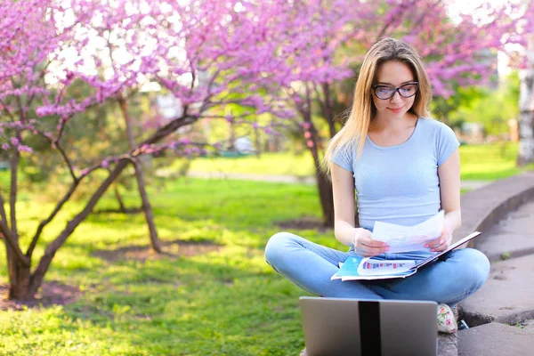 Étudiante étudiant avec ordinateur portable et papiers dans le parc en fleurs . — Photo