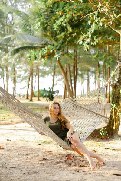 Молодая женщина сидит босиком на плетеном гамаке, песок и деревья на заднем плане . — стоковое фото