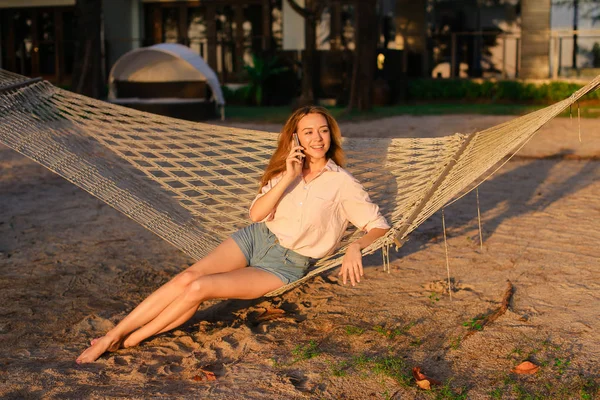 Hamakta oturmuş ve smartphone, yaz tatillerini konuşan genç beyaz kadın.