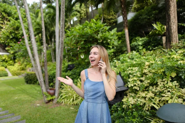 Jovem mulher conversando por smartphone no jardim perto de árvores, vestindo jeans sundress . — Fotografia de Stock