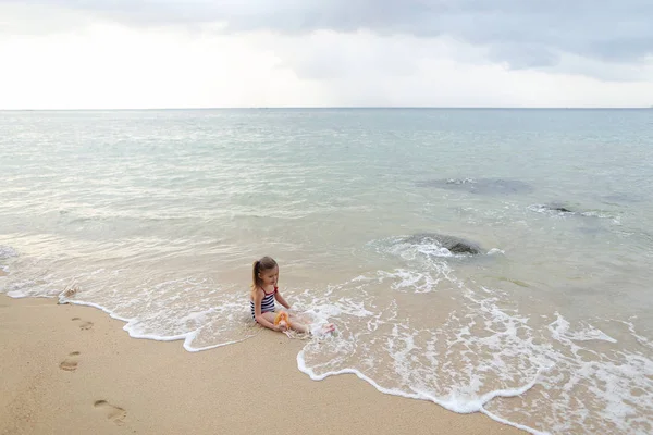 海と遊ぶ人形の近くの砂の上に座っている少女. — ストック写真