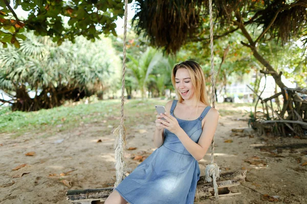 Молодая красивая женщина катается на качелях и используя смартфон, песок и дерево на заднем плане . — стоковое фото