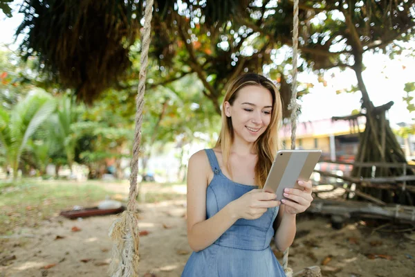 Chica joven usando la tableta y montar columpio en la arena, usando pantalones vaqueros vestido . — Foto de Stock