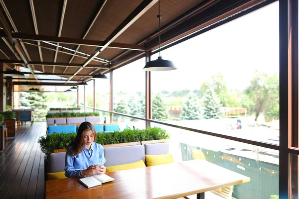 Chica caucásica sentada en la cafetería con teléfono inteligente y anotando en el diario . — Foto de Stock