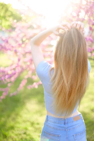 Blond flicka stående i solen strålar med grönt gräs och blossom bakgrund. — Stockfoto