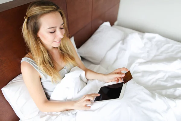 Молода біла жінка бронює квитки онлайн за планшетною та золотою дебетовою карткою, сидячи в ліжку . — стокове фото