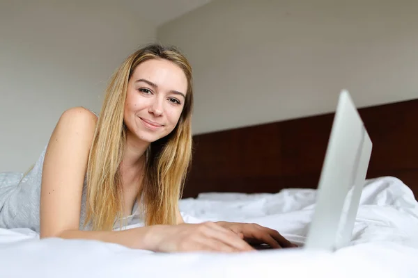 Joven mujer feliz usando el ordenador portátil y acostado en la cama por la mañana . — Foto de Stock