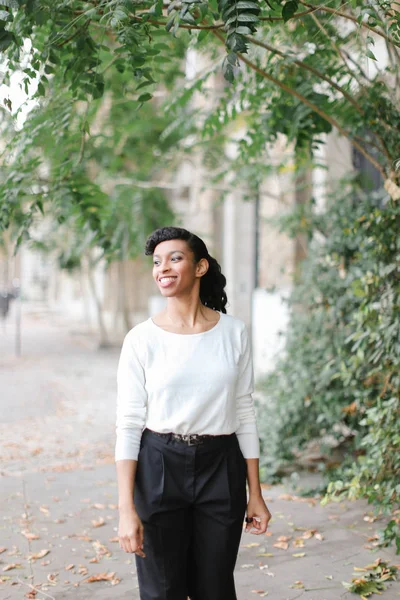 Black smiling female person walking near building and green trees, wearing white blouse and black pants.