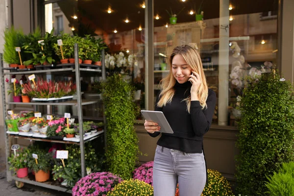 Mujer joven gerente de floristería de pie con la tableta y hablando por teléfono inteligente . — Foto de Stock