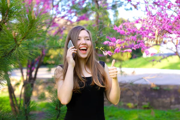 Mädchen spricht mit Smartphone und zeigt Daumen nach oben im Frühlingspark. — Stockfoto