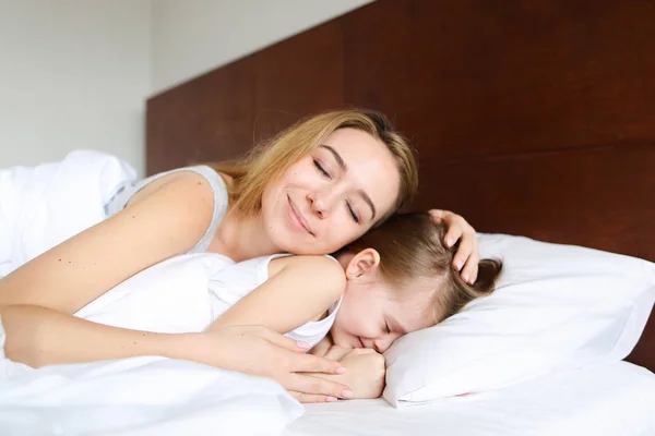 Jovem mulher bonita dormindo com a pequena filha de manhã na cama com lençóis brancos . — Fotografia de Stock