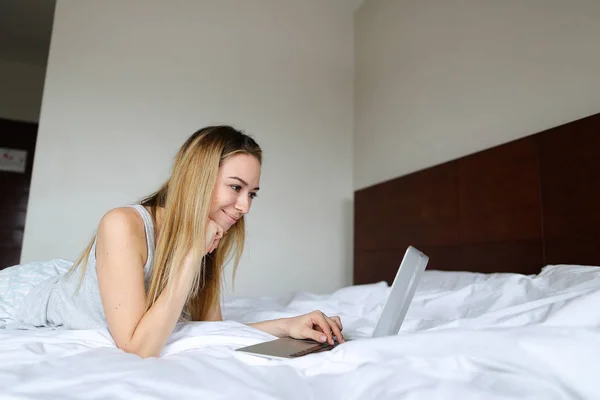 Chica rubia joven usando el ordenador portátil y acostado en la cama por la mañana . — Foto de Stock