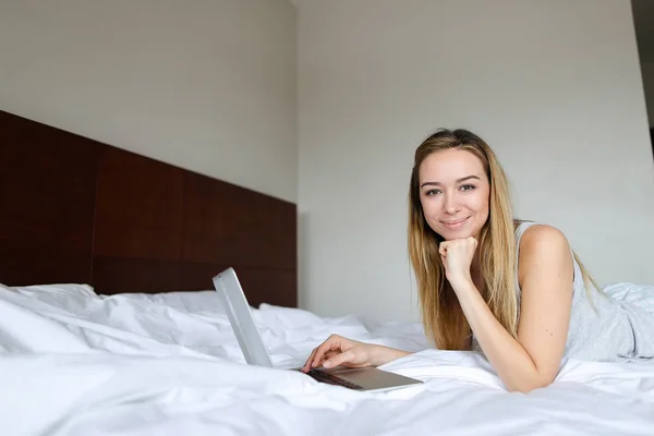Joven chica encantadora usando el ordenador portátil y acostado en la cama por la mañana . — Foto de Stock