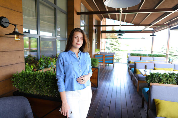 Pretty woman standing at cafe with smartphone and wearing jeans shirt and white pants.