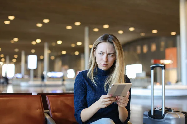 Caucasian female person sitting in airport waiting room with valise and browsing by tablet.