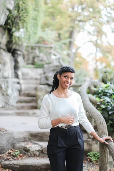 Happy black girl wearing white blouse standing near stone stairs in park.