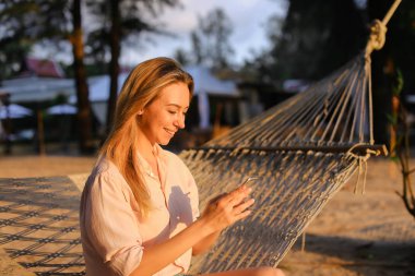 Smartphone kullanarak ve hasır hamak tropikal resort üzerinde oturan genç Amerikalı kadın.