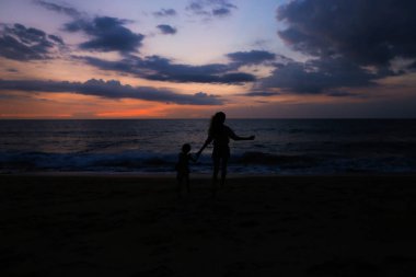 Bir anne ile küçük kız deniz kıyısında gündoğumu arka planda çalışan siyah siluet.