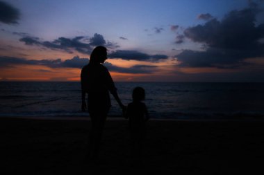 Anne kızın elini tutarak ve gün batımı artalan ile deniz kıyısında yürüyen siyah silüeti.