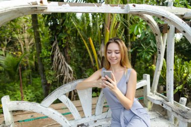 Salıncakta sürme ve egzotik Bahçe, avuç içi arka plan smartphone kullanarak genç güzel kadın.