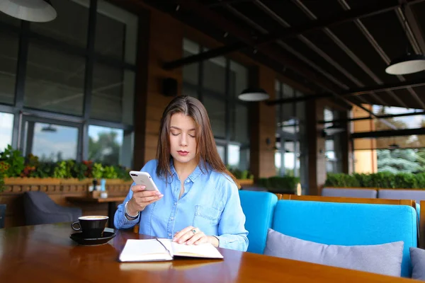 Linda chica leyendo diario cerca de la taza de café en la cafetería y el uso de teléfono inteligente . — Foto de Stock