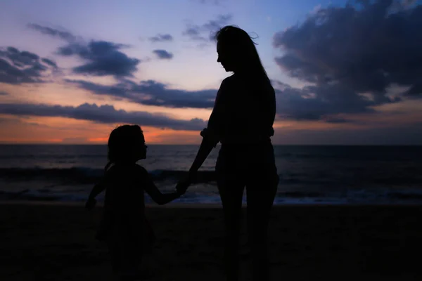 夕日を背景に海の近くの小さな女の子と歩く若い女性人の黒いシルエット. — ストック写真