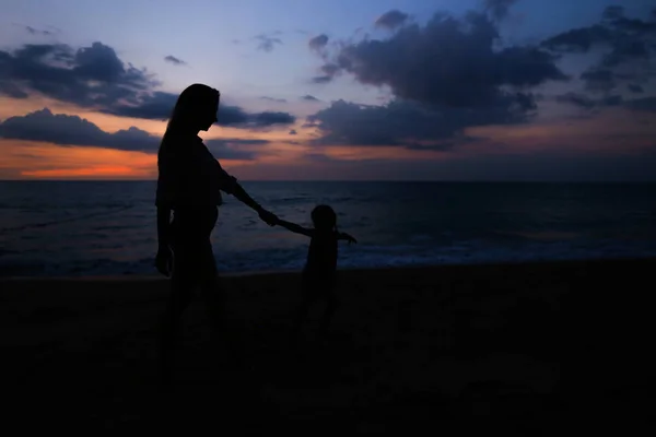 Sziluettjét anya lánya kezemet, és a séta a tenger partján naplementében háttérbeli. — Stock Fotó