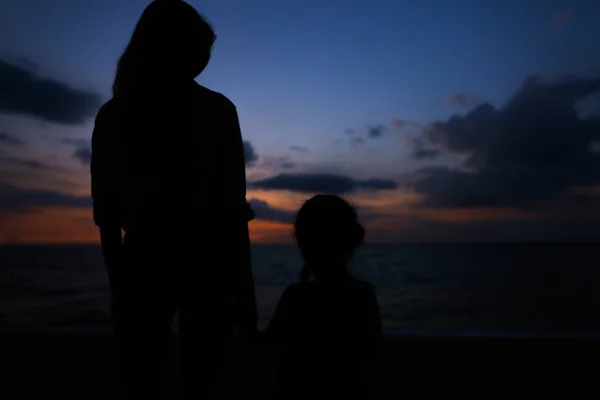 Black silhouette of female person holding daughter hand and walking on sea shore with sunset backgound. — Stock Photo, Image