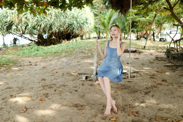 Unga europeiska kvinna Rider swing och tal av smatrphone, sand och träd i bakgrunden. — Stockfoto