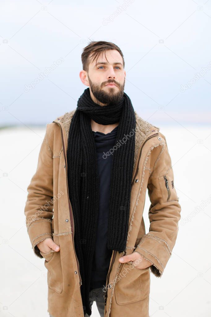 European young male person with beard wearing coat and scarf standing in white snow background.