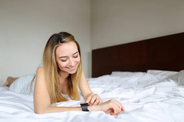 Joven bonita mujer usando smartwatch y acostado en la cama con ropa de cama blanca en la mañana . — Foto de Stock