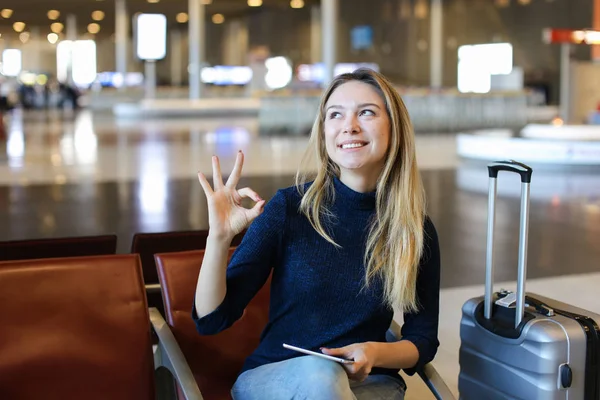 Dziewczyna siedzi w poczekalni Lotnisko z valise, używając tabletu i pokazano ok. — Zdjęcie stockowe