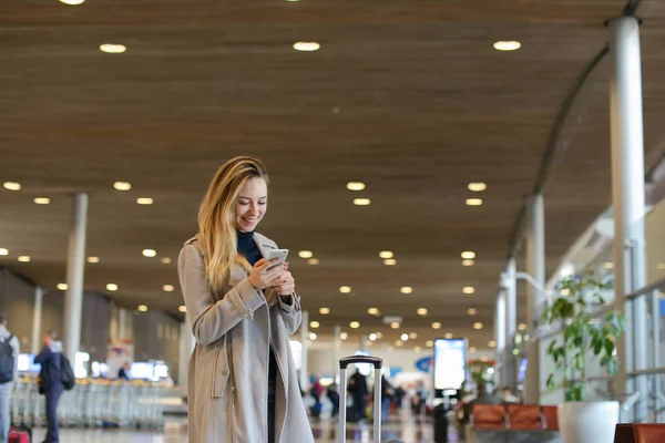 Joven mujer en la sala del aeropuerto de pie con valise y el uso de teléfono inteligente . — Foto de Stock